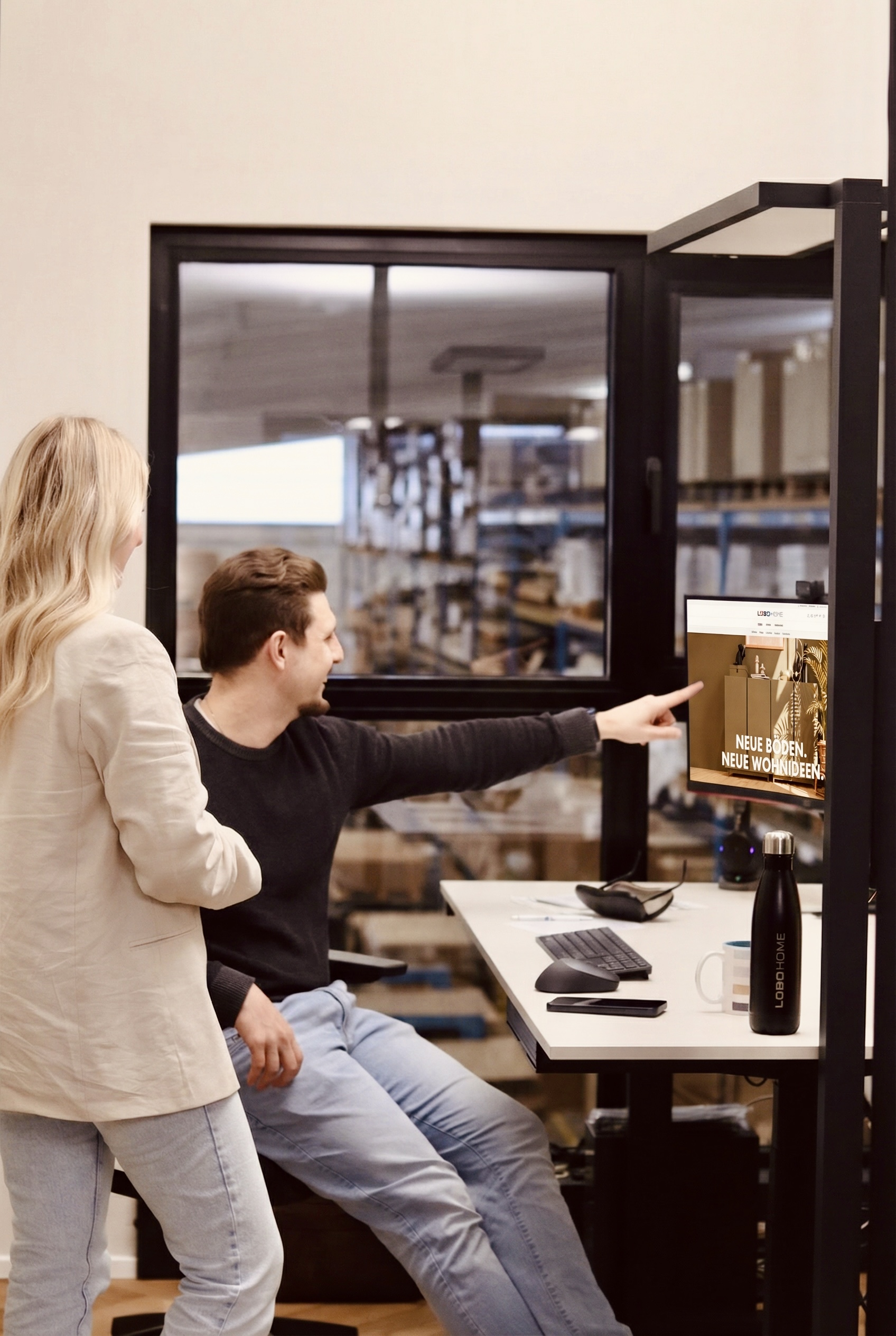 Zwei Mitarbeitende von LOBO HOME betrachten gemeinsam den Online-Shop an einem Arbeitsplatz mit Blick ins Lager